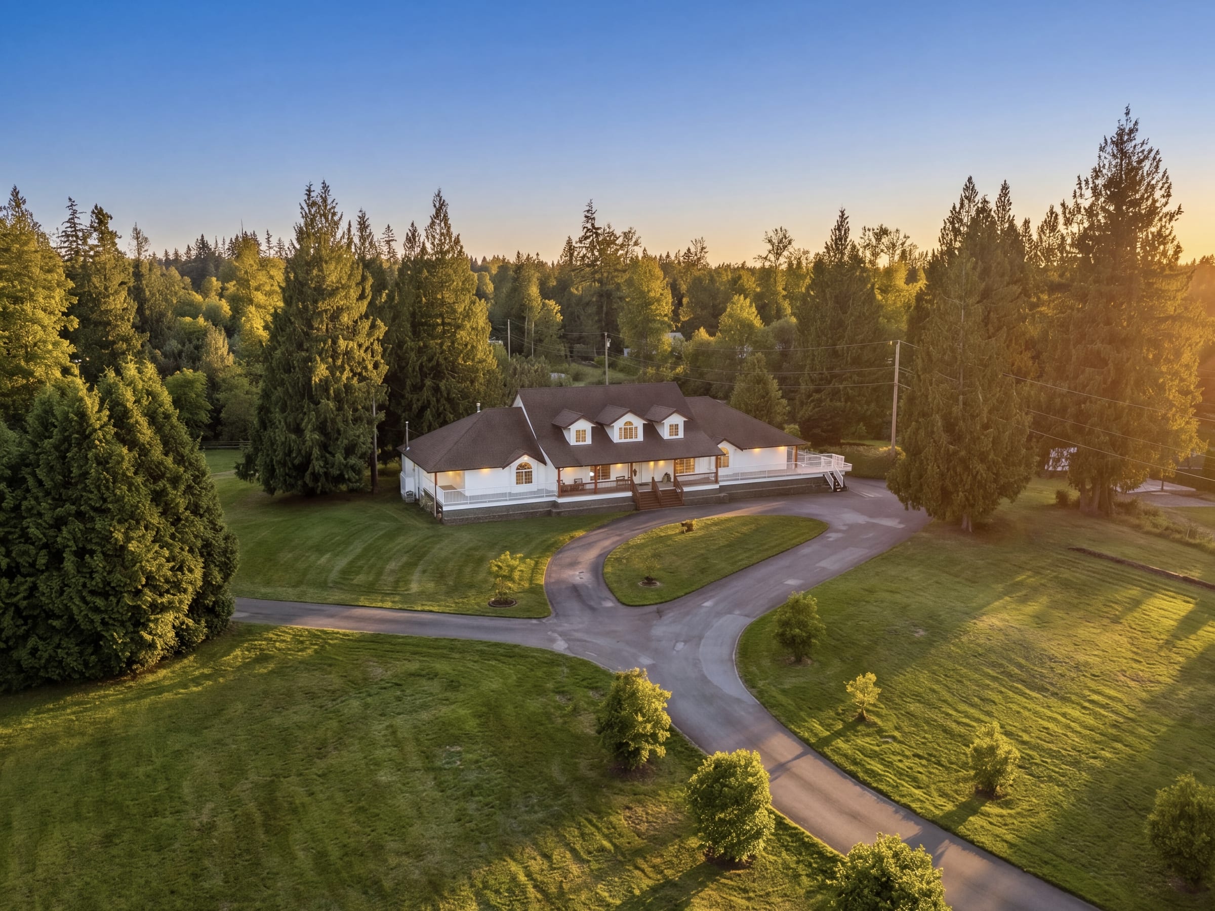 21980 100 Avenue in Fort Langley: a white rancher with dormers on 4.2 acres, a circular drive and tall evergreens, at dusk