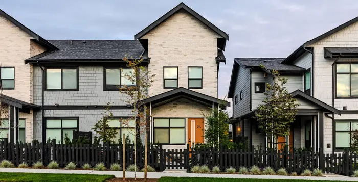 Photograph of the finished Gordon Square townhomes: brick and pale siding with black window frames and an orange front door, behind a dark picket fence with young trees
