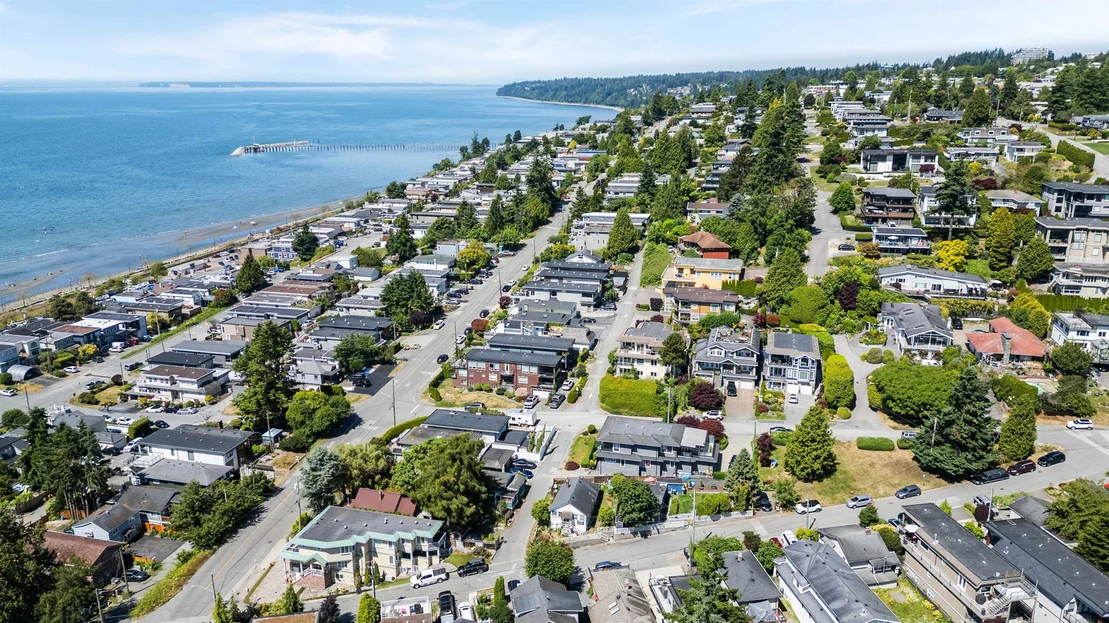 Aerial view of the South Surrey / White Rock waterfront — the curved beach, the White Rock pier extending into Semiahmoo Bay, residential blocks climbing the hillside toward the trees