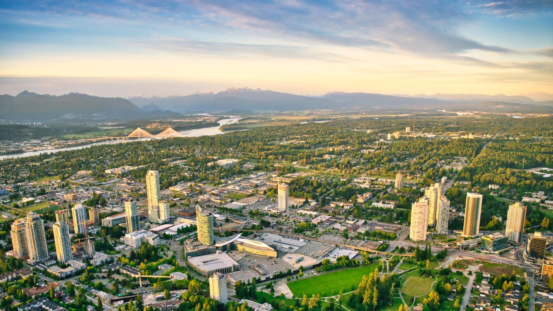 Aerial view of Surrey City Centre at golden hour — concrete towers around Surrey Central and King George stations, Holland Park in the foreground, the Fraser River and Pattullo Bridge in the distance toward the North Shore mountains