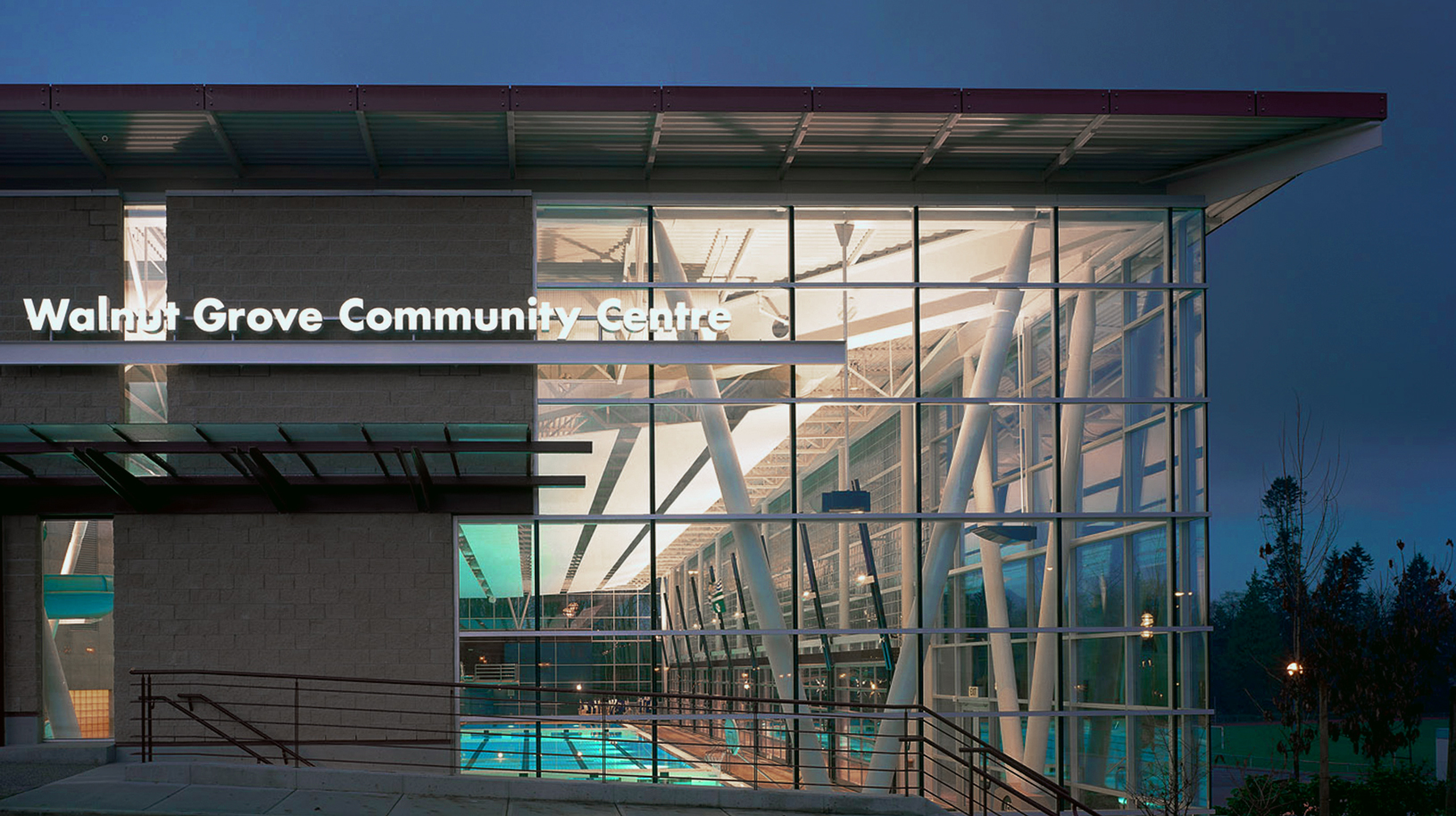Walnut Grove Community Centre at dusk — the curved-glass aquatic-centre wing with the village name on the canopy, lit interior visible through the floor-to-ceiling glass