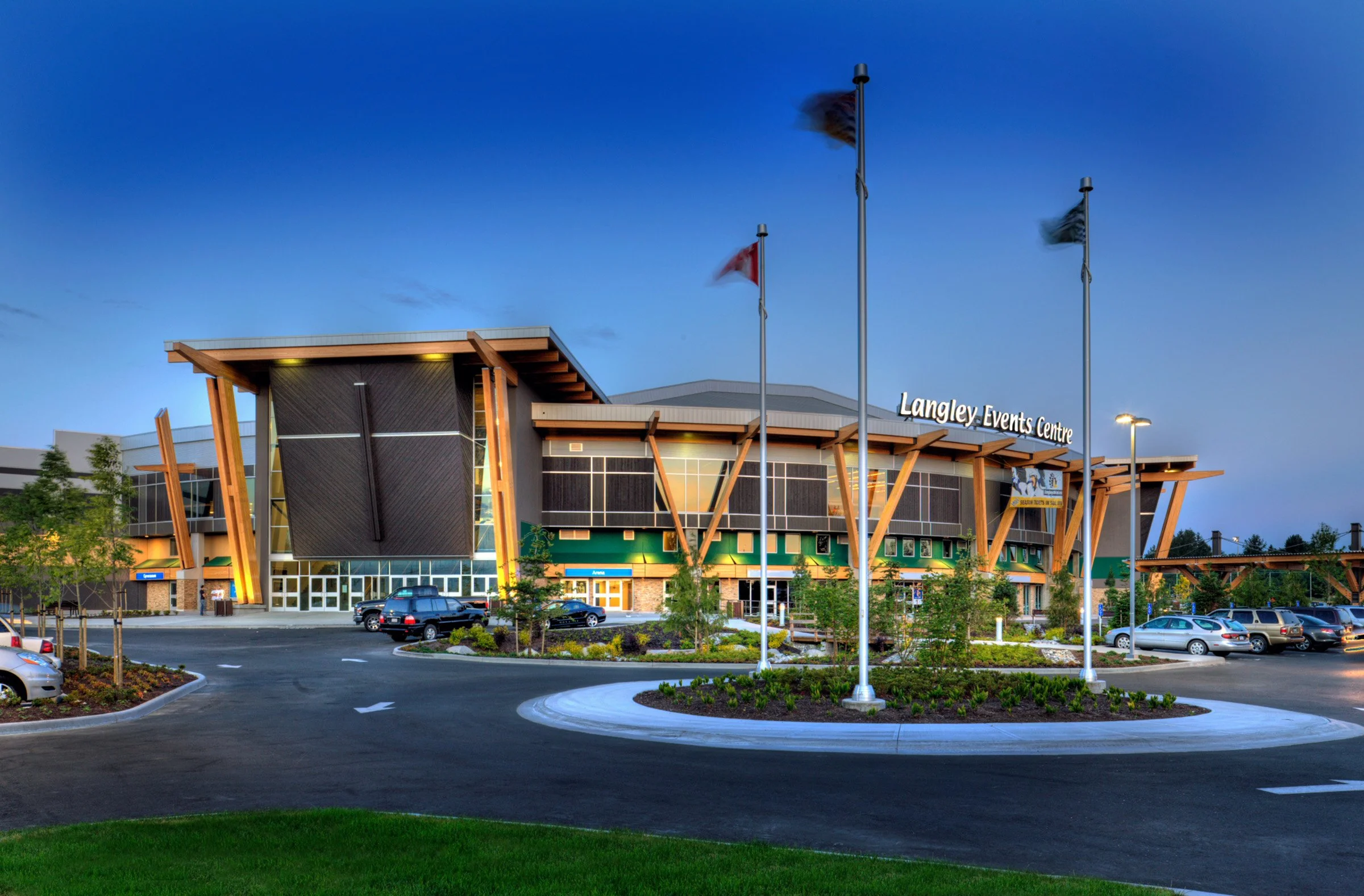 Langley Events Centre at dusk — the heavy-timber peaked entrance with flagpoles at the roundabout drop-off, anchor of the 200 Street commercial spine